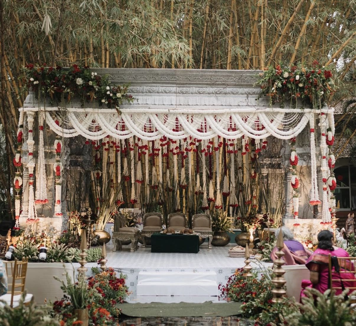 Wedding mandap draped in strands of white jasmine and red roses, set against a bamboo grove